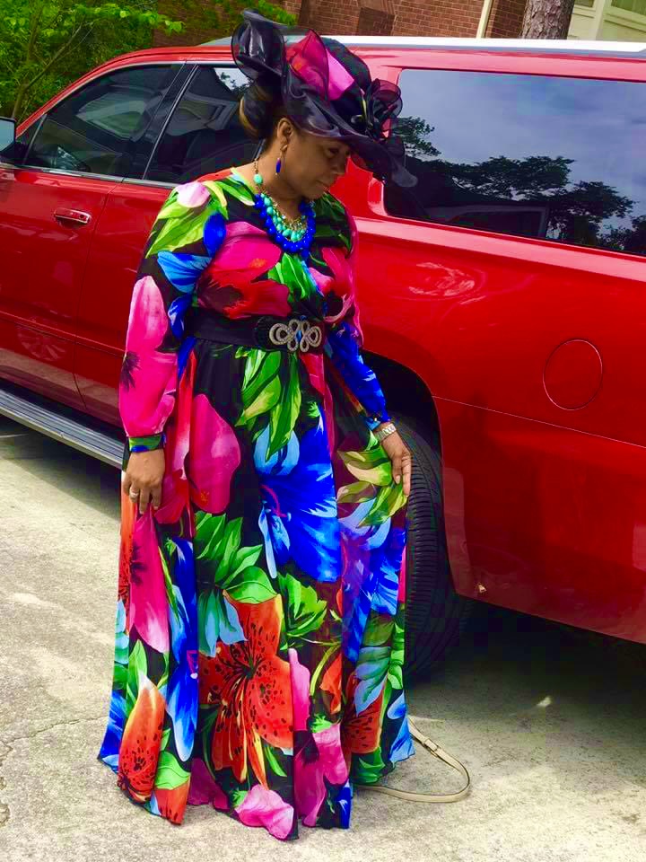 Mary Kingcannon in a vibrant floral dress and statement hat beside a red SUV
